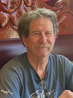 A smiling man with a mustache, wearing a blue shirt and sunglasses hanging from his neck, seated in front of a decorative wall.
