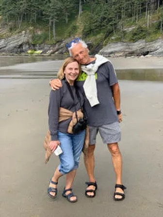 A smiling couple embracing on a sandy beach with cliffs and trees in the background.