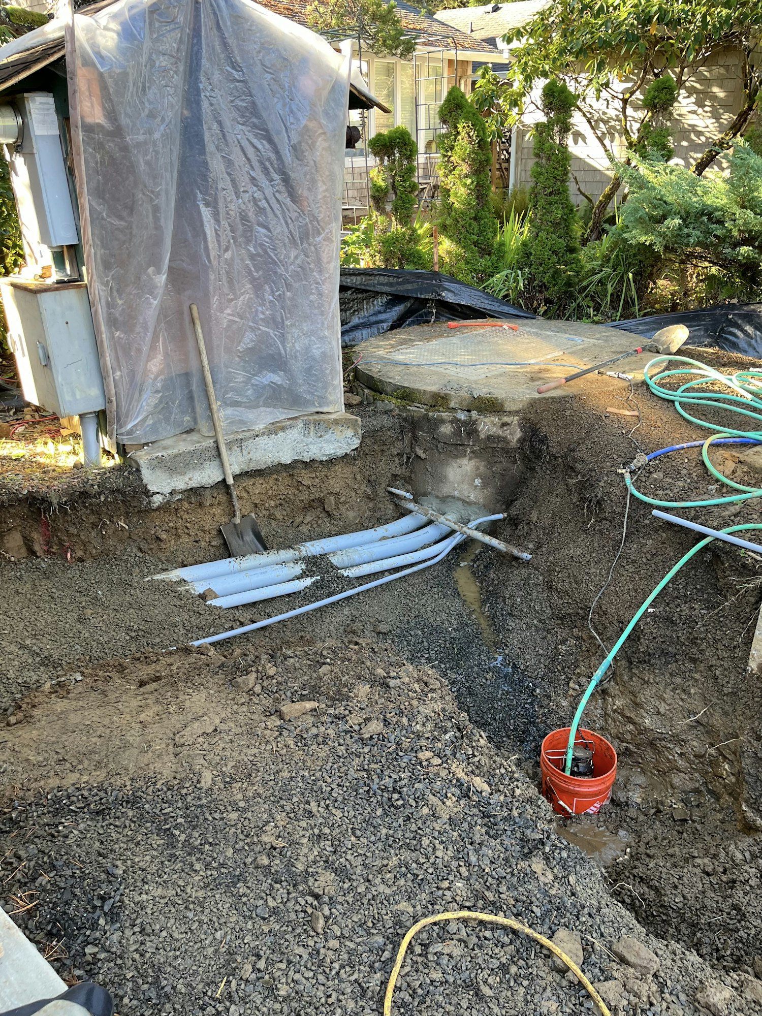 A dug-up area with exposed pipes, a shovel, a bucket, and hoses near a house, likely indicating construction or repair work.