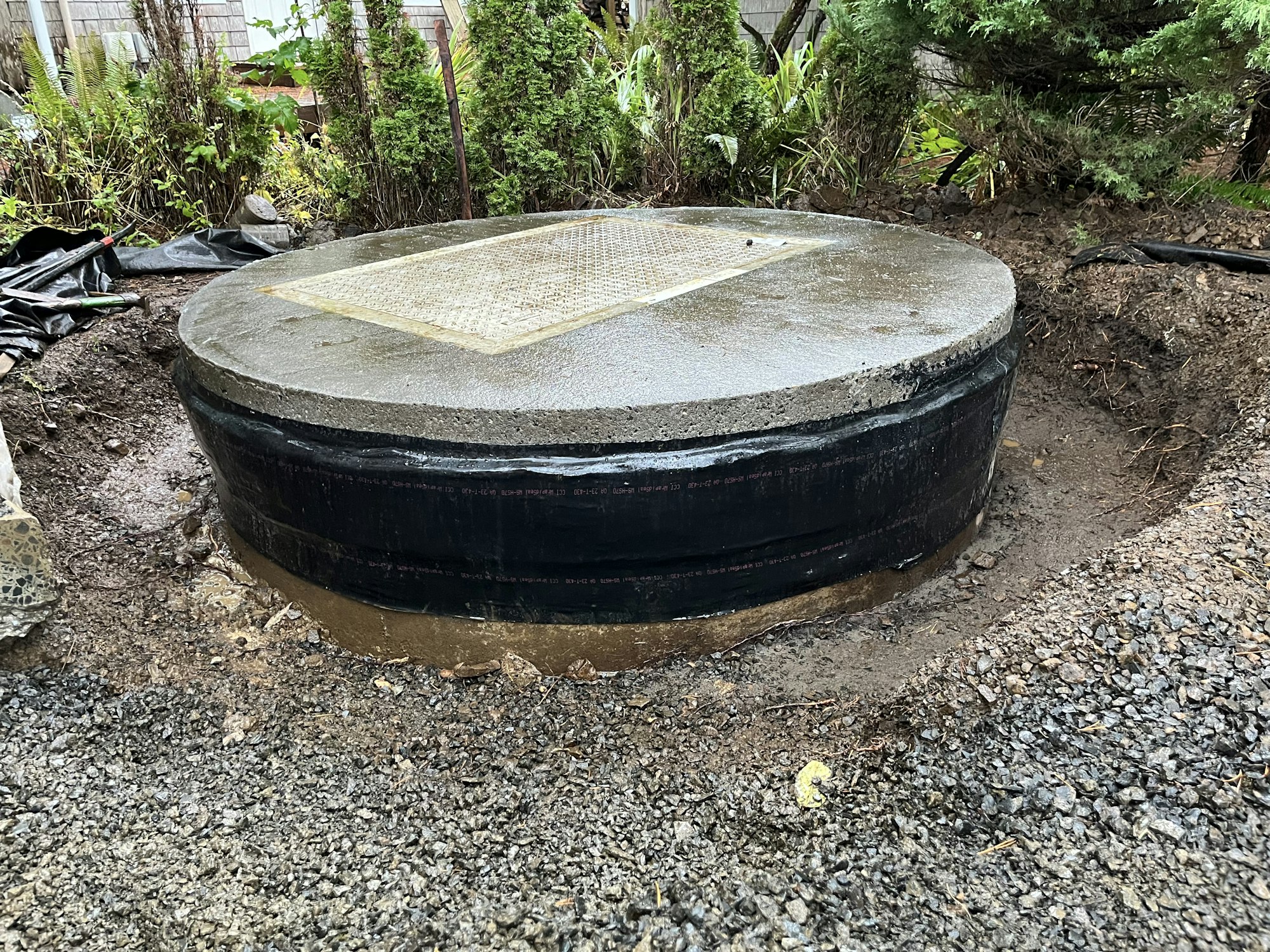 A concrete septic tank lid on a freshly installed or serviced tank, surrounded by dug-up earth and garden foliage.