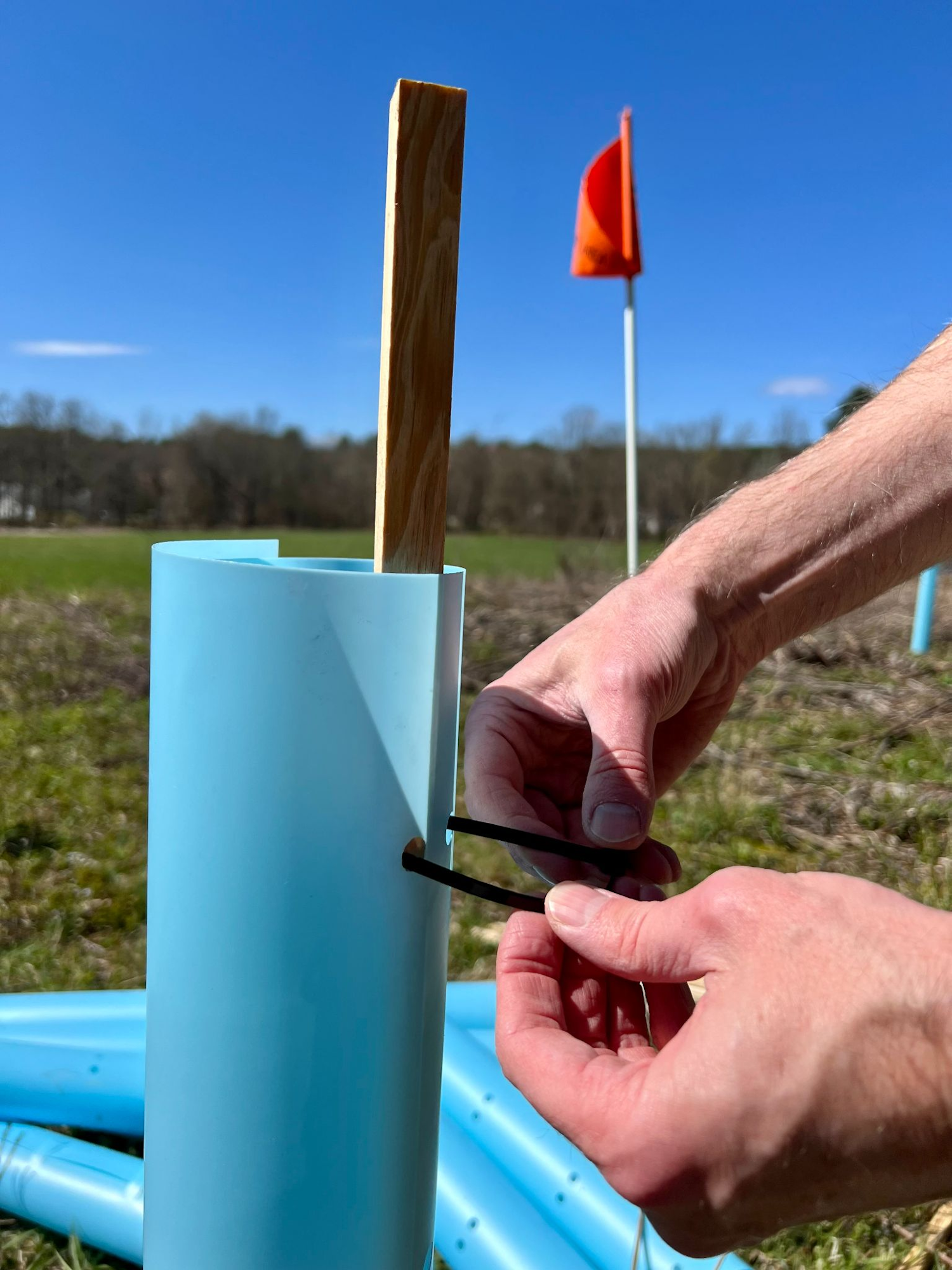 A person securing a stake with a cotter pin in a blue cylinder, outdoor setting, with an orange flag in the background.