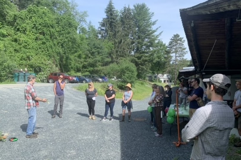 A group of people gathered outdoors, possibly for an event or meeting, with greenery and cars in the background.