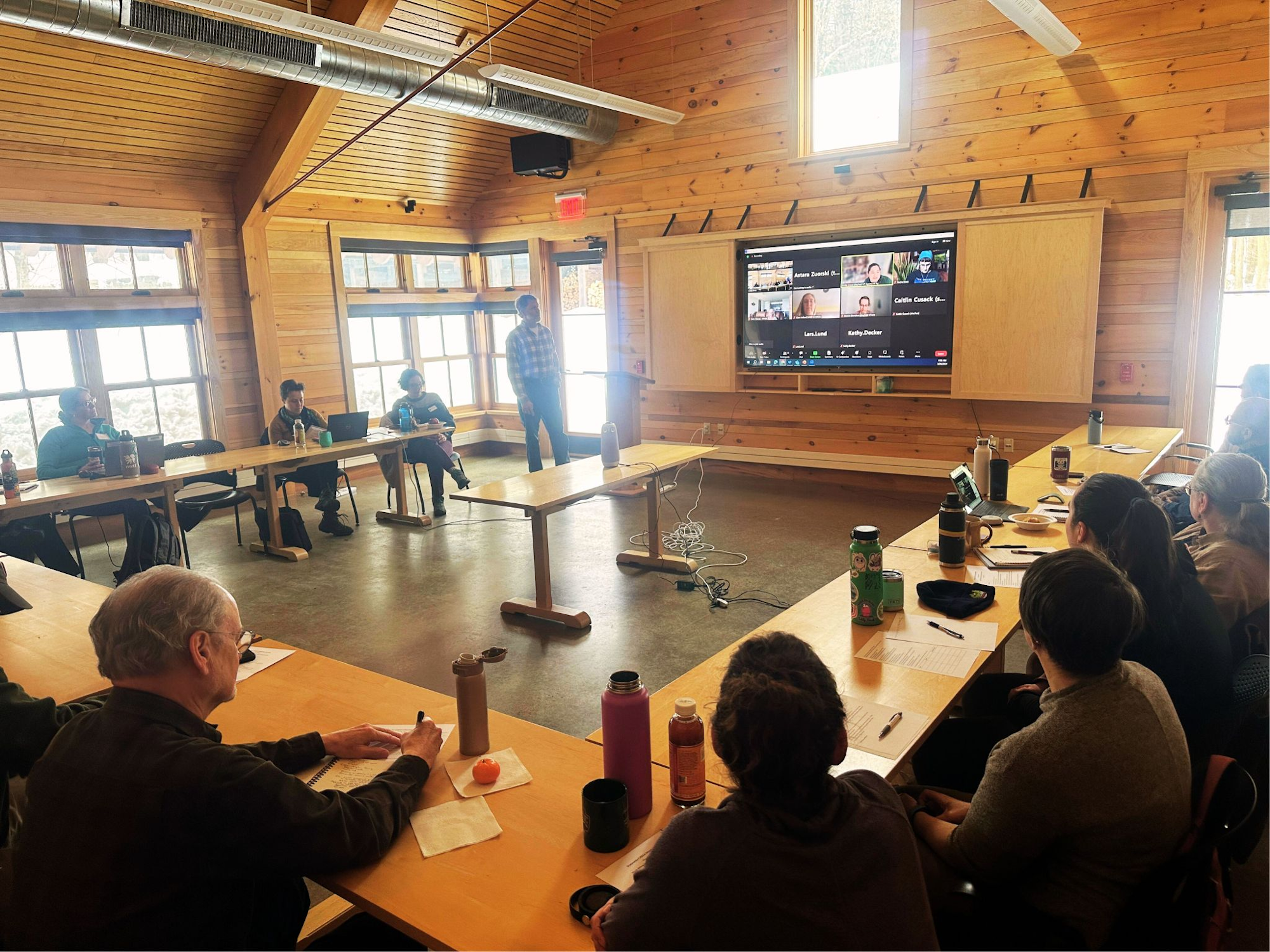 A room with people at tables facing a screen with a video conference, suggesting a hybrid meeting or workshop.