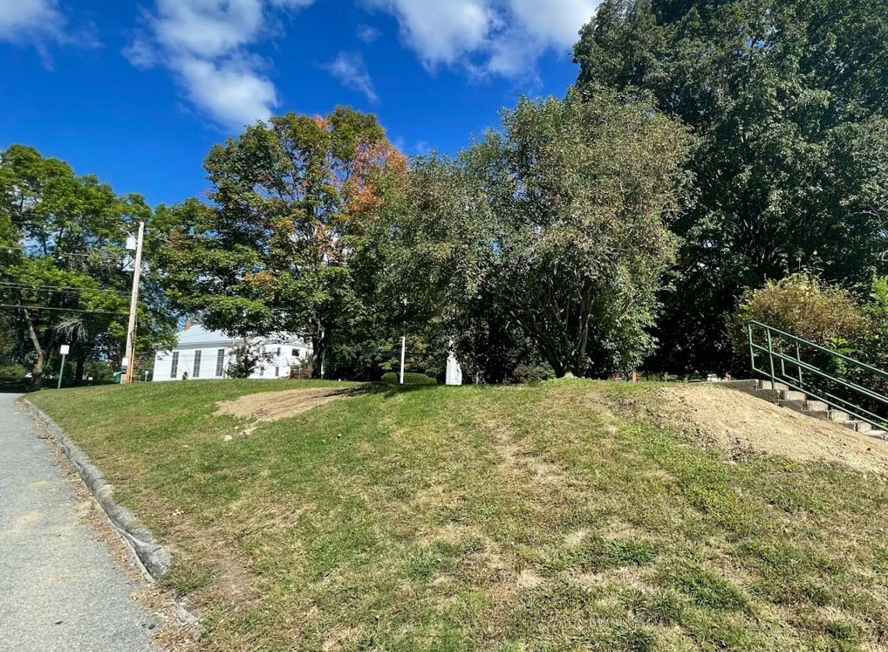 A grassy slope with trees, a path, railing, and a white building under a blue sky with clouds.