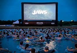 Outdoor movie screening of "JAWS" with audience floating in water on inflatables at dusk.