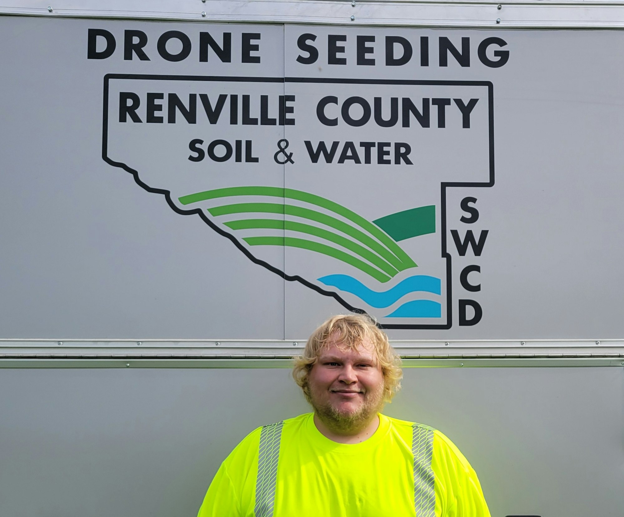 A person in a bright yellow shirt stands in front of a "Renville County Soil & Water" sign about drone seeding.