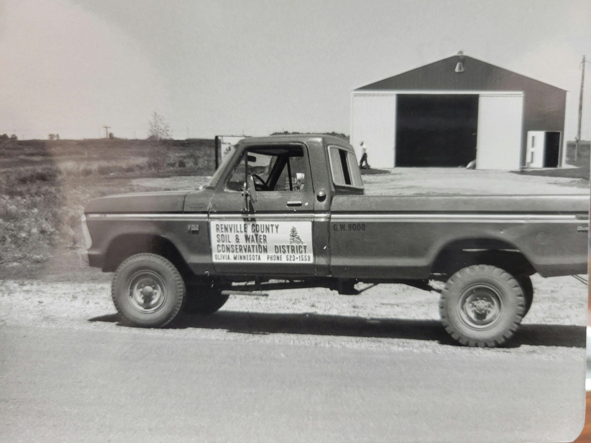 Vintage photo of a Renville County Soil & Water Conservation District pick-up truck outside a garage, with a person in the background.