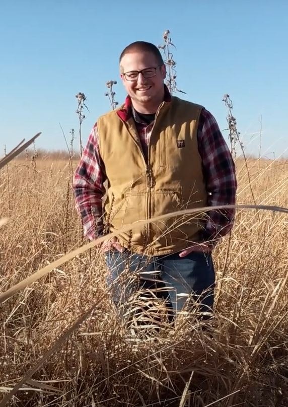 Kyle Richter, smiling, in a vest and plaid shirt standing in a field with tall, dry grass under a blue sky.