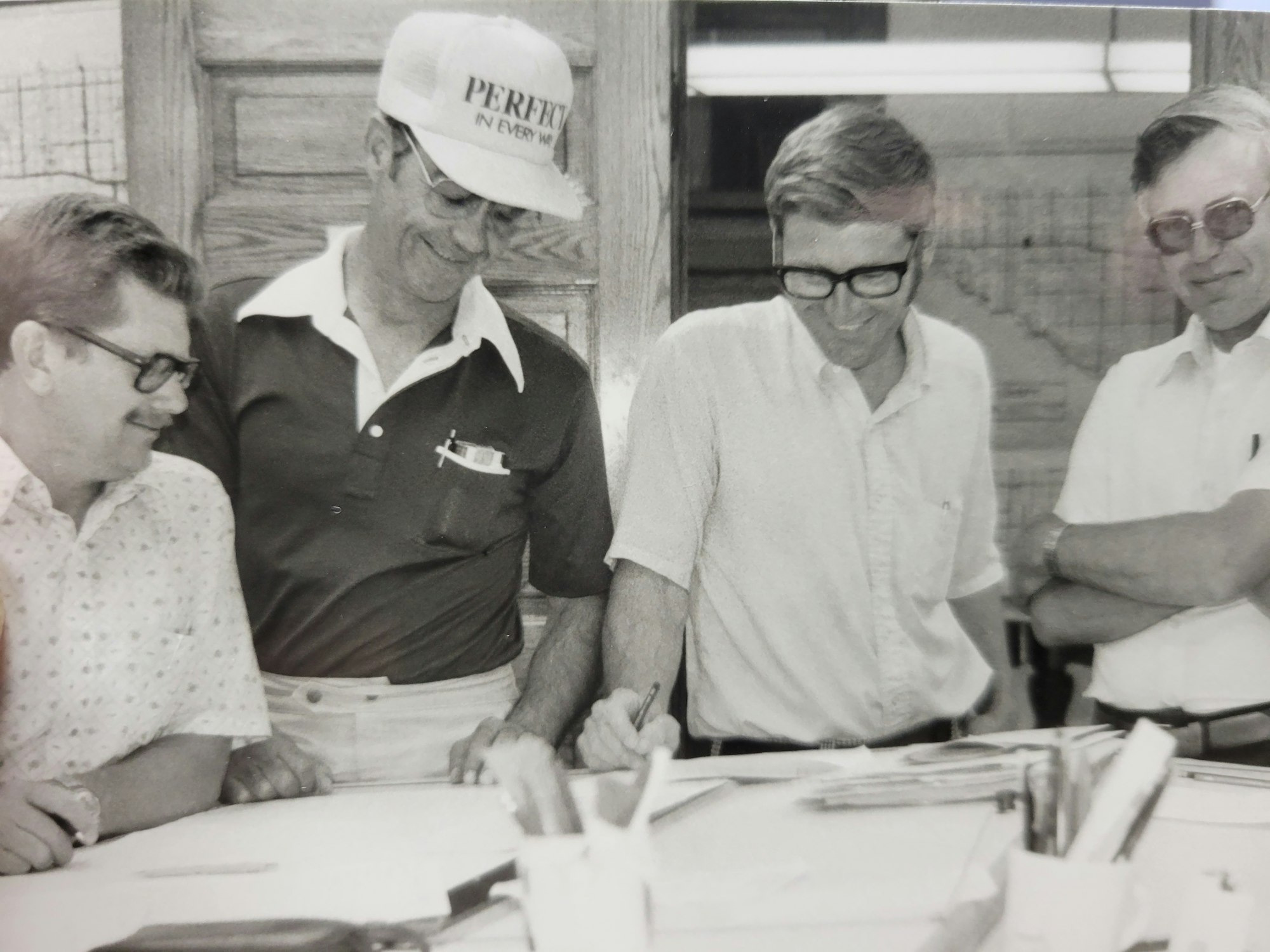 Four men reviewing papers, smiling, casual attire, indoors, vintage black and white photo.