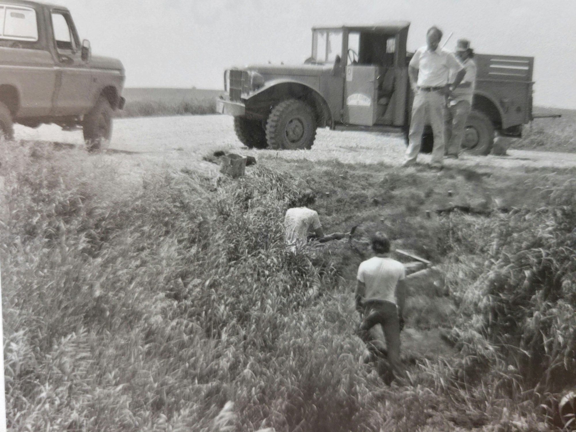 Two trucks and three people, one digging, in a field.