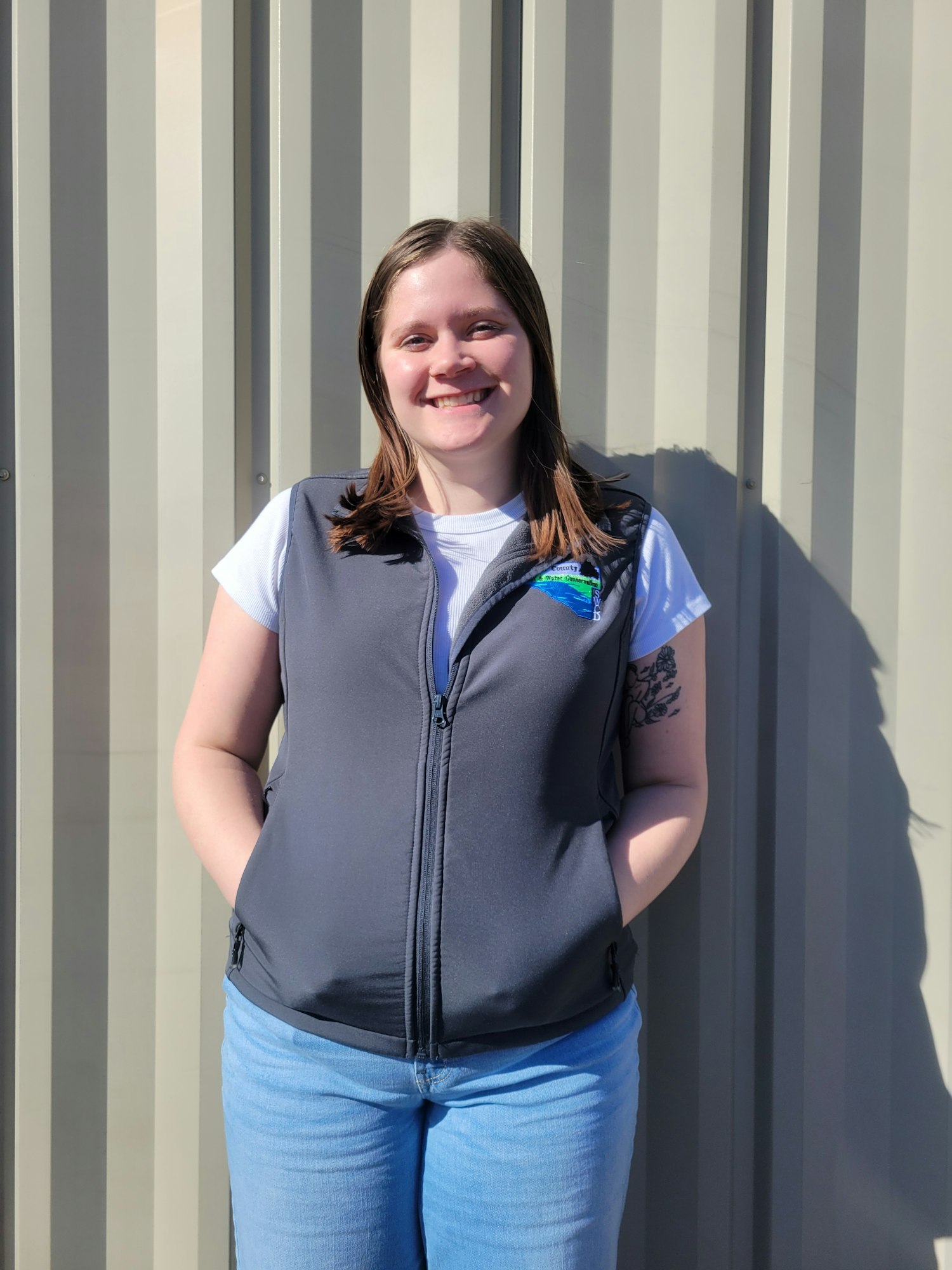 Pamela Winzenburg, smiling, standing by a metal wall, wearing a white shirt and a grey Renville SWCD vest.