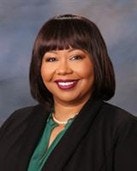 A person smiling, wearing a dark suit jacket, green shirt, and pearl necklace against a neutral background.