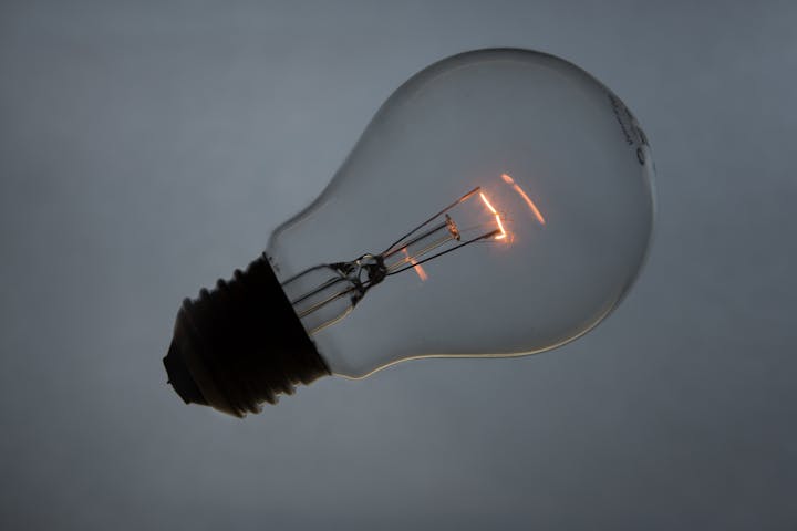 A glowing light bulb, emitting a warm light, against a grey background, highlighting its filament inside.