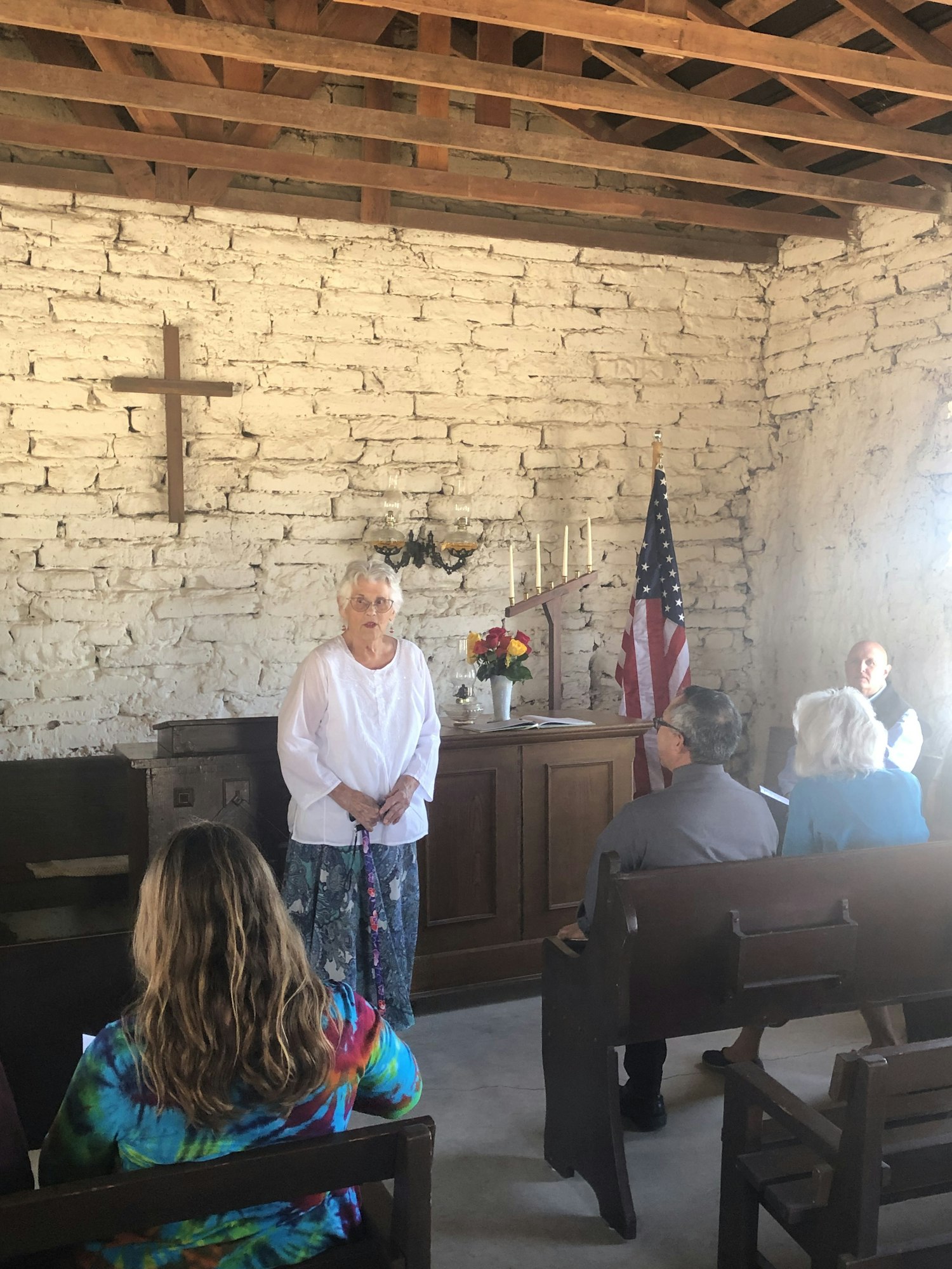 A small group gathers in a rustic room with a U.S. flag, cross on the wall, and an altar. A person stands at the front speaking.