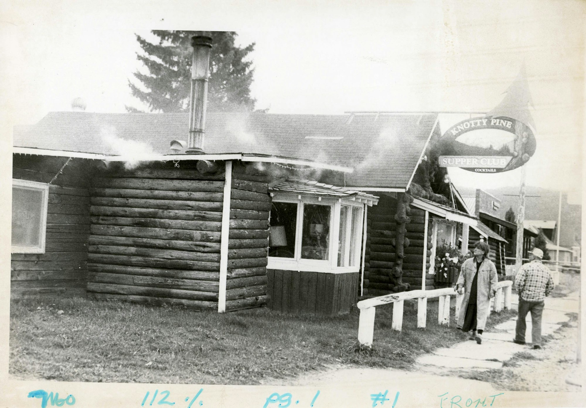 Black and white photo of a log cabin-style building with a sign "Knotty Pine Supper Club," smoke coming out, and people outside.