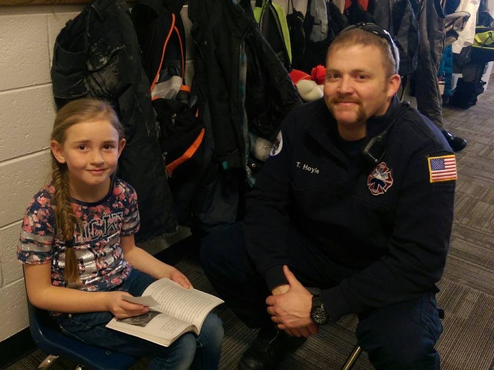 A girl reading a book next to a firefighter with a thumbs-up, coats in the background.