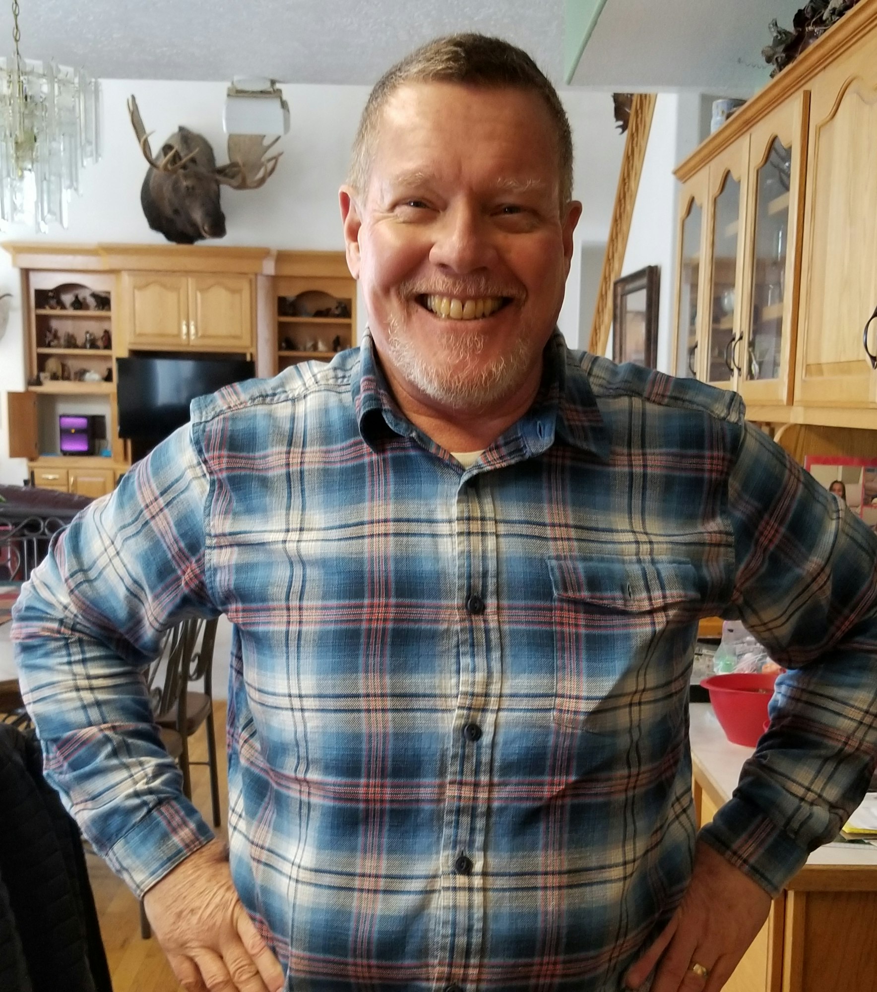 A smiling man in a plaid shirt stands in a kitchen with cabinetry and a moose head on the wall.