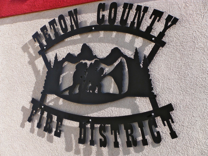 Metal sign for "Teton County Fire District" with silhouette of firefighters and mountains.