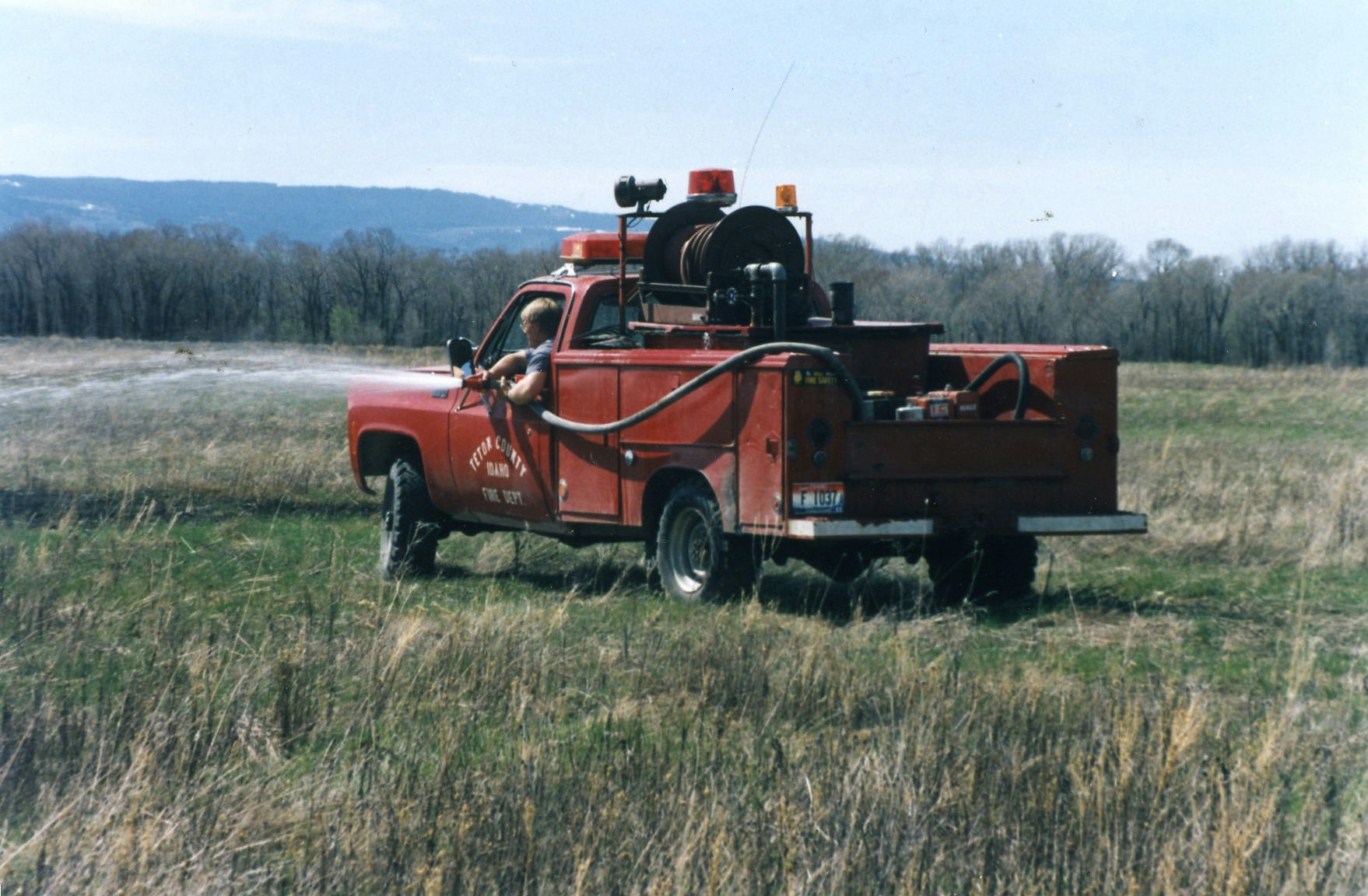 A red utility truck with equipment and a person inside is parked in a grassy field.