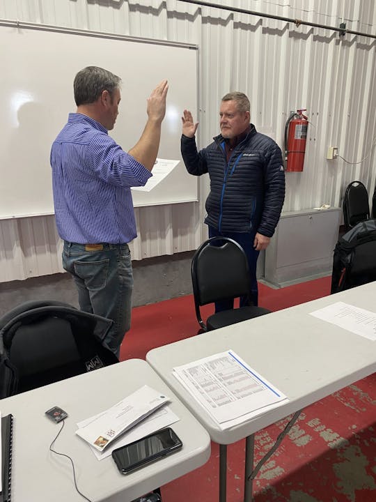 Two men standing, one with raised hand, by a whiteboard and table inside a room.