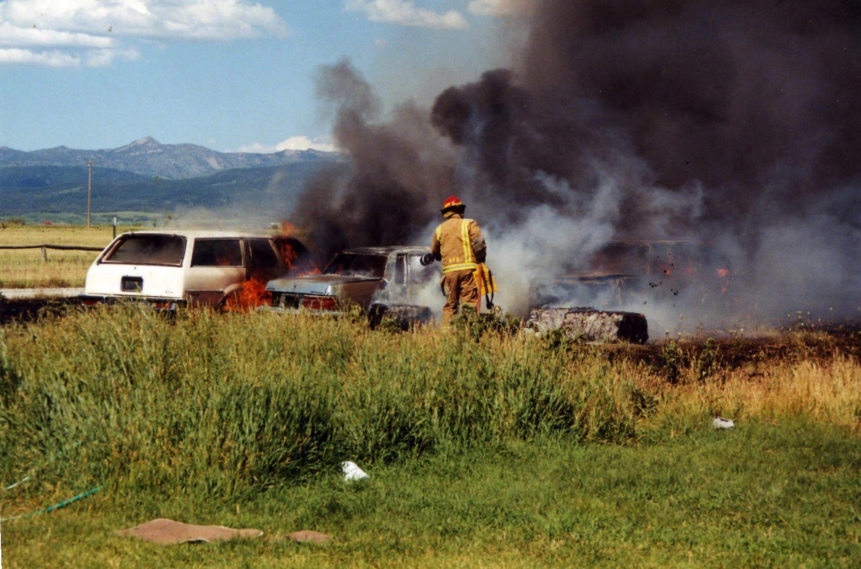 A firefighter battles a blaze engulfing two cars in a grassy field with mountains in the background.