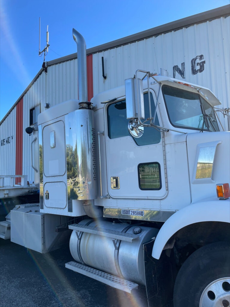 Part of a white semi-truck with exhaust stack and fuel tank, parked near a building with "ING" sign.