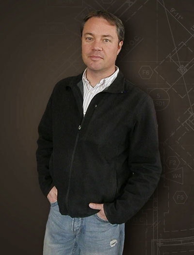 A man in a black jacket and blue jeans standing before a background with technical drawings.
