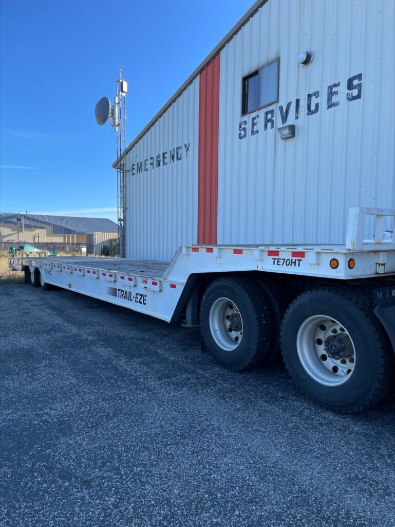 A flatbed trailer in front of a building with "EMERGENCY SERVICES" sign and an antenna on a clear day.