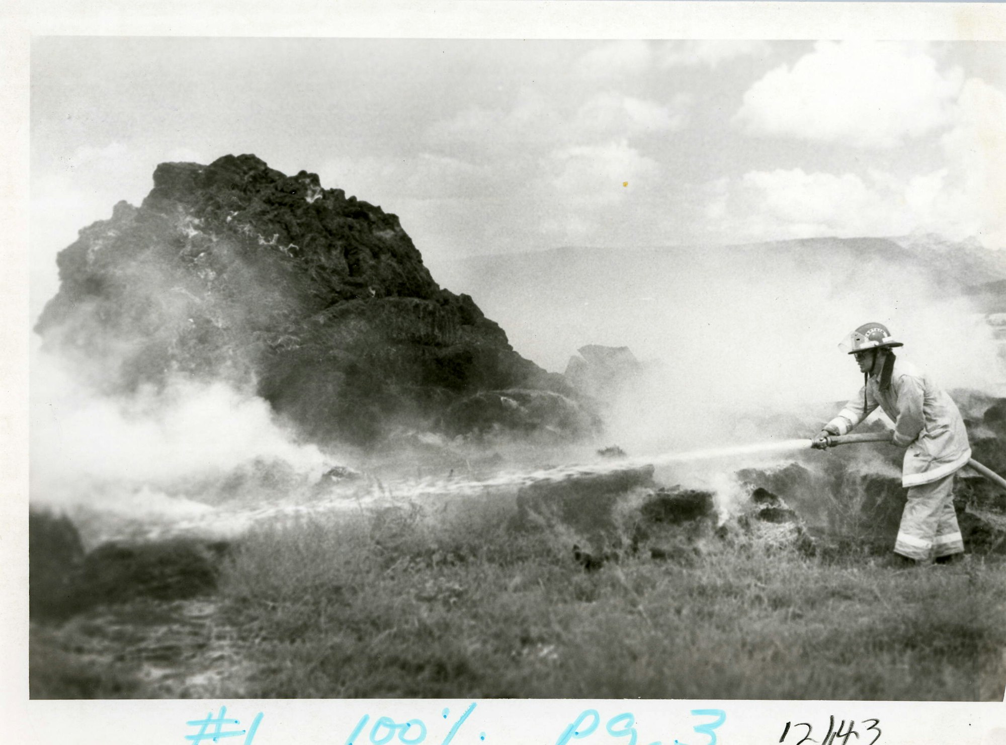 A person in a helmet and coat using a hose, possibly firefighting, against a smoky background with rocky terrain.