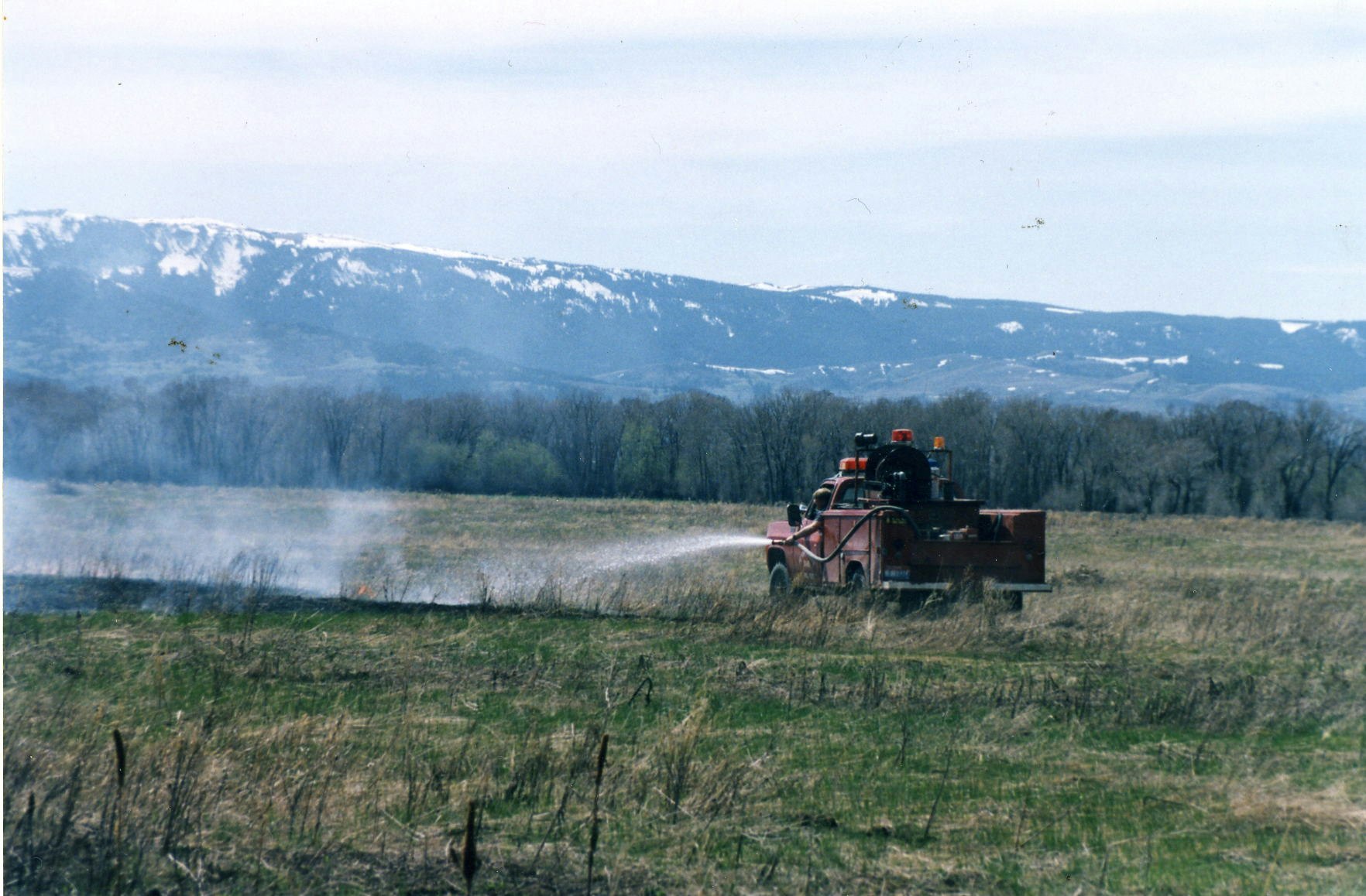 Firefighting truck in a field with a smoke plume, mountains in the background.