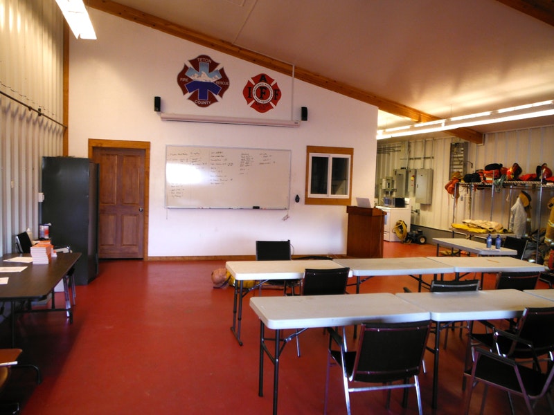 Interior of a fire station training room with tables, chairs, whiteboard, and firefighting gear.