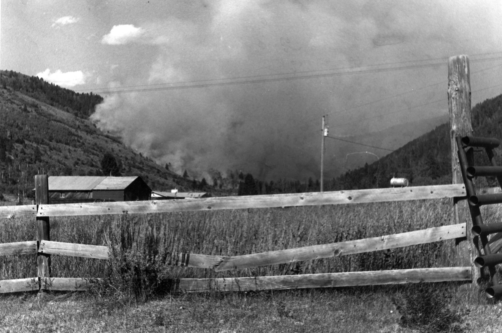 A black and white photo showing a wooden fence, a barn, and smoke from a distant wildfire in a mountainous area.