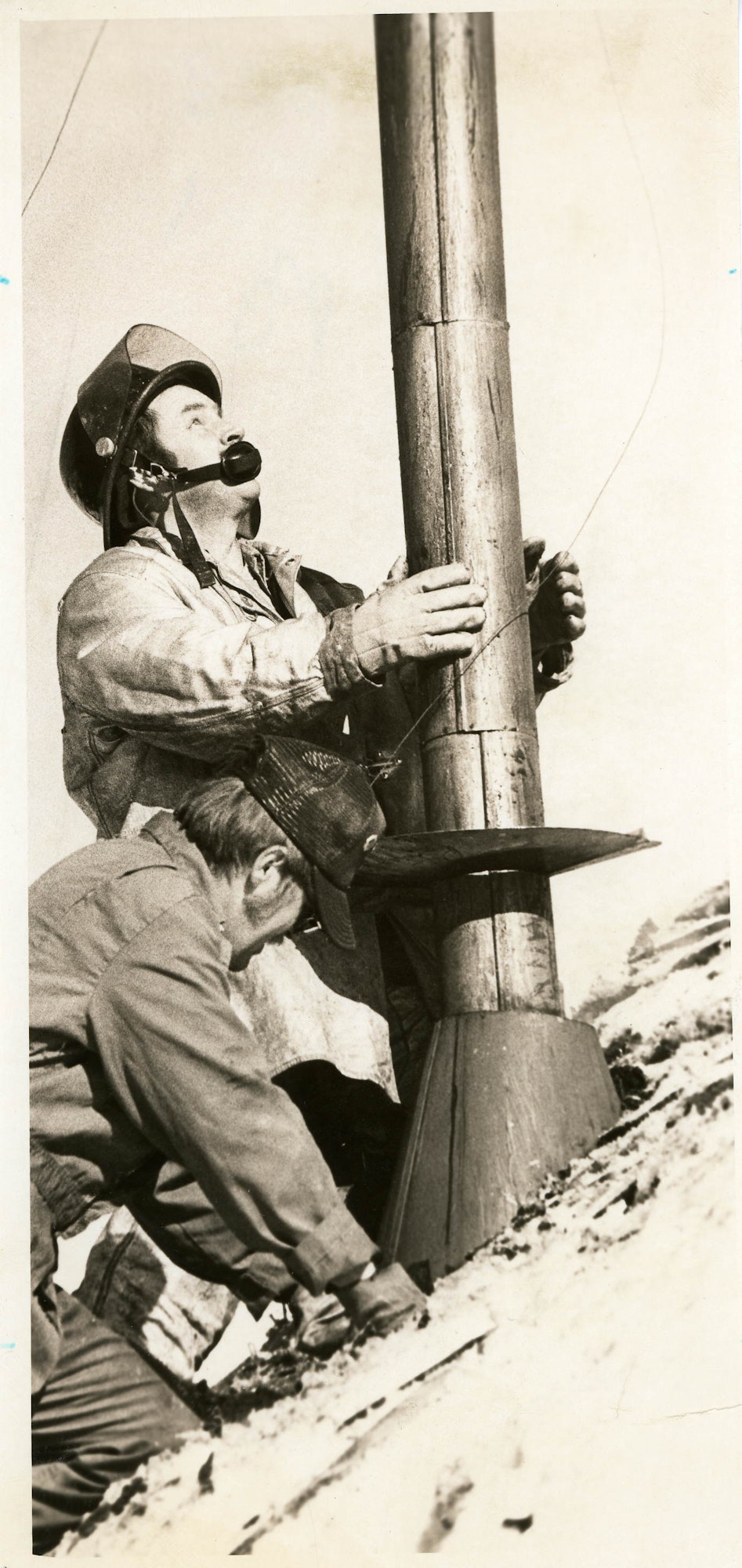 Two men installing or repairing a telephone pole, one standing with a headset, the other crouched below. Vintage photograph.