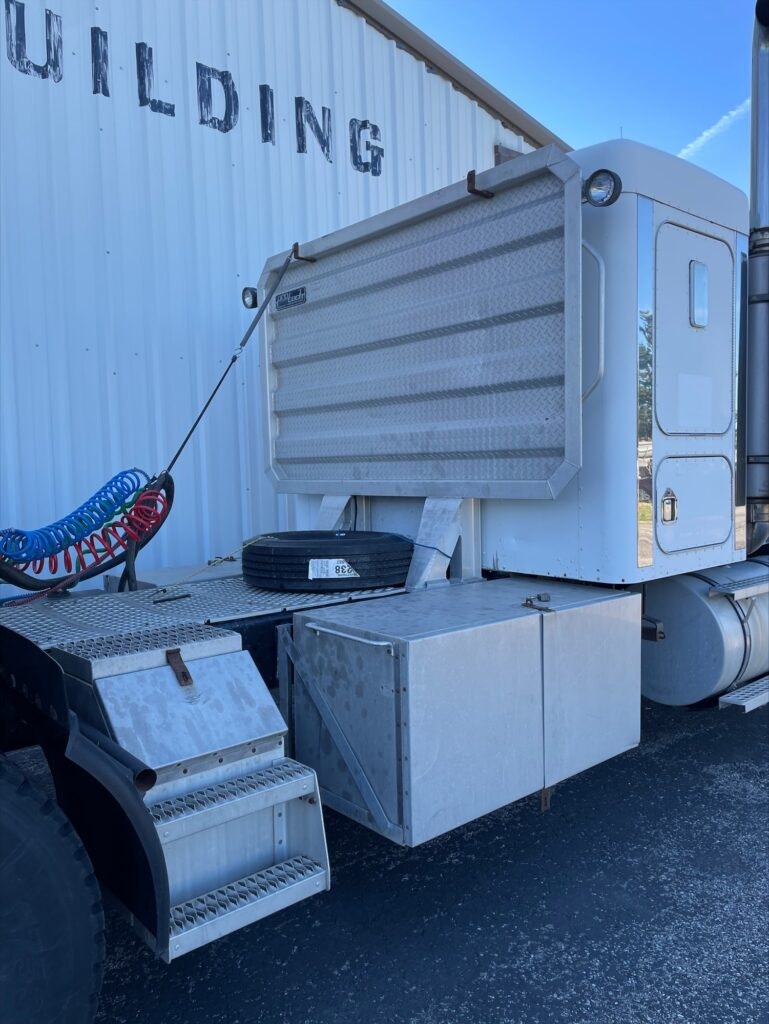 Semi-truck with open storage compartments, a coiled air hose, tire, and part of a building.