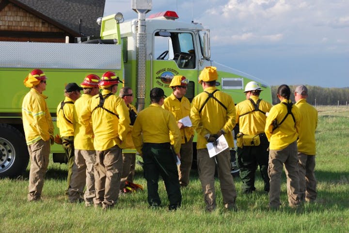 Group of firefighters in yellow uniforms beside a firetruck, possibly in a briefing or debriefing.