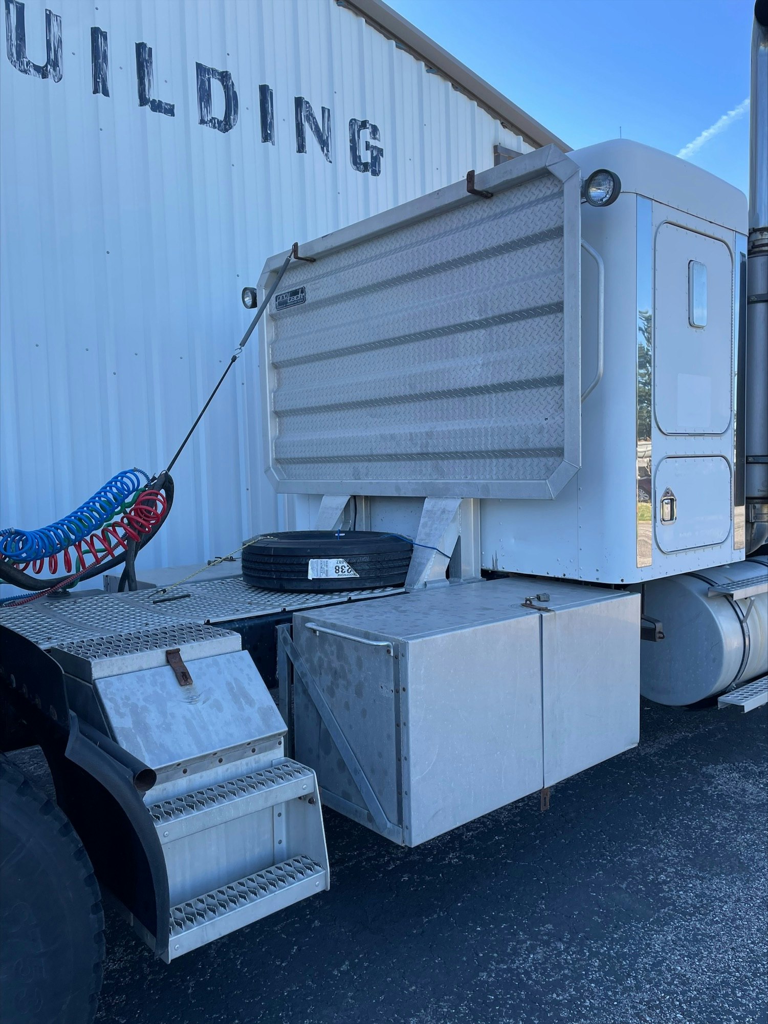 Semi-truck with open side storage compartment and steps, parked by a building, under a clear sky.