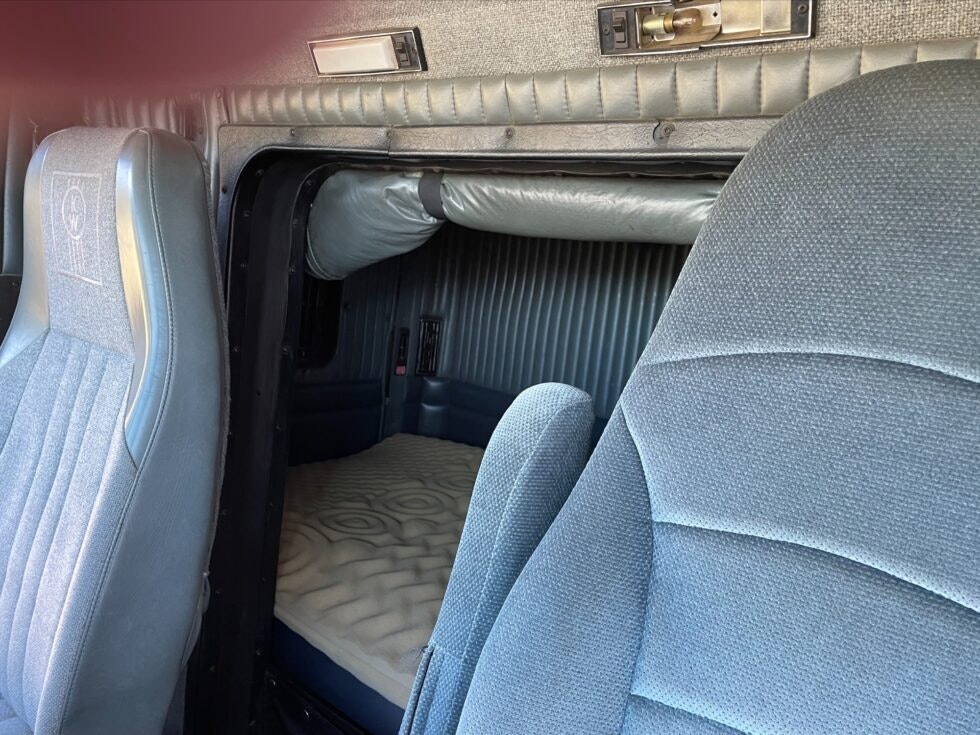 Interior view of a vehicle, showing part of front seats and a view into the sleeper compartment with a bed.