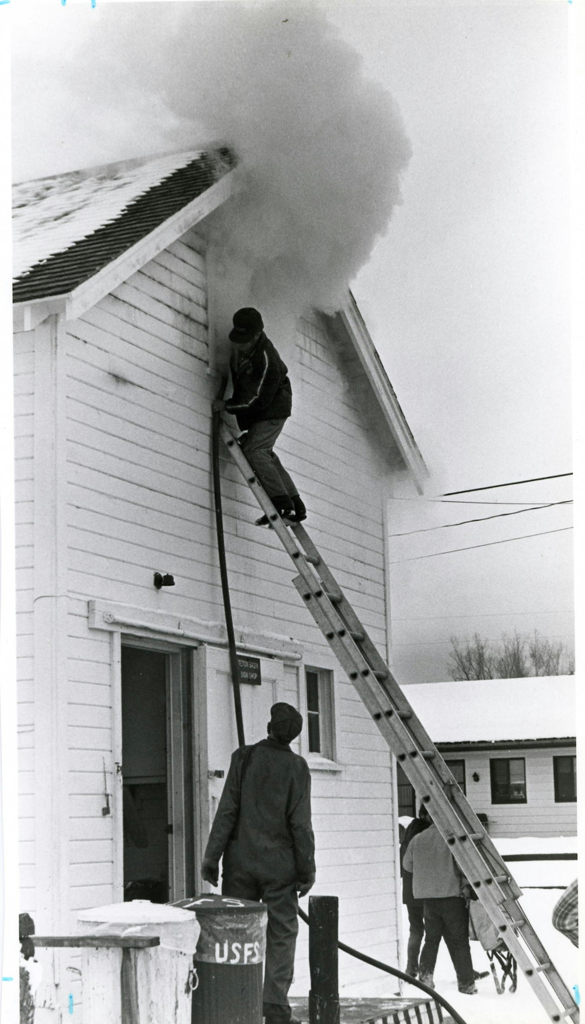 Person on ladder by a smoking chimney; another person watches. Cold weather attire suggests wintertime.