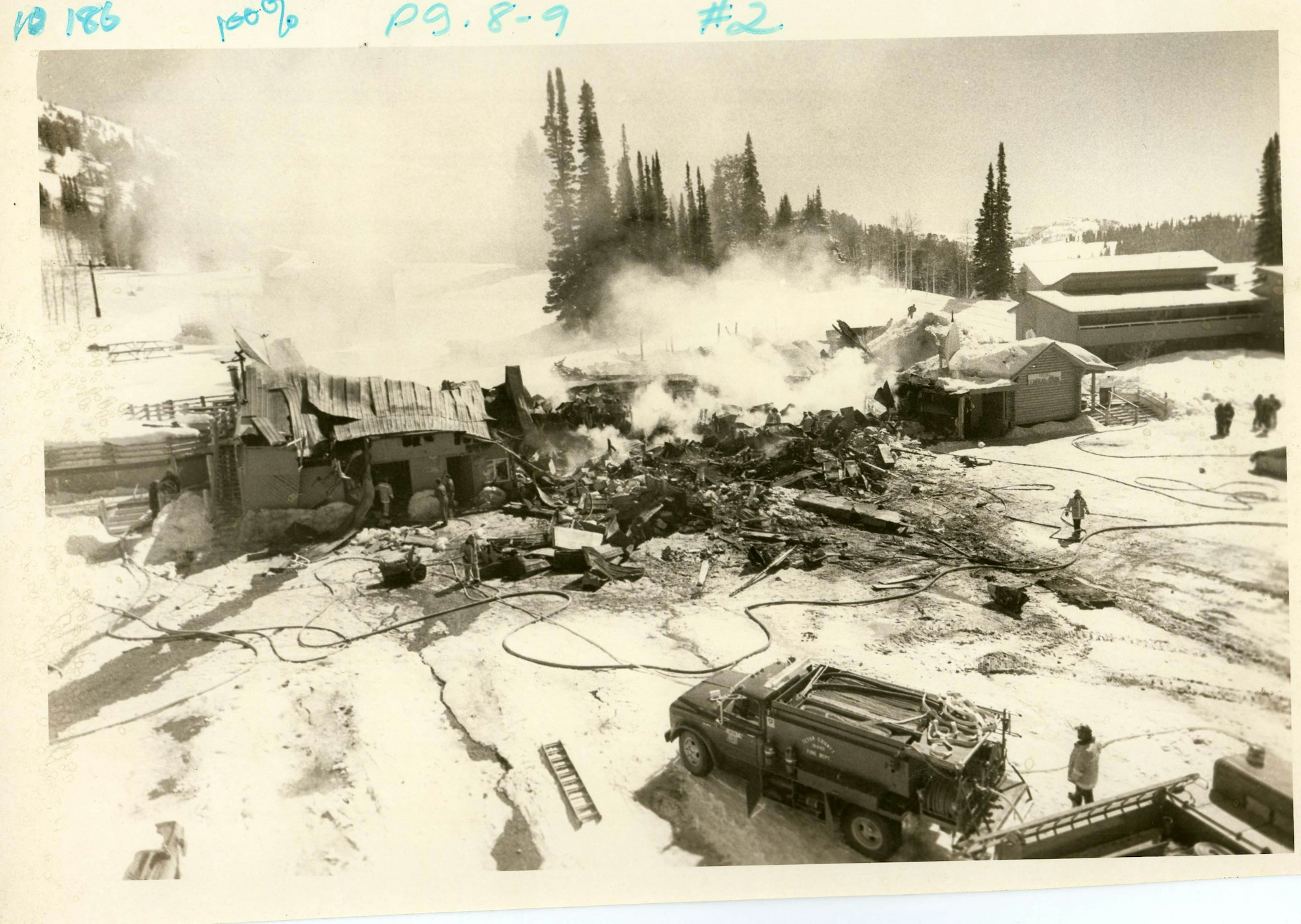 A black and white photo of a collapsed building with firefighters, smoke, and emergency vehicles amidst snowy surroundings.