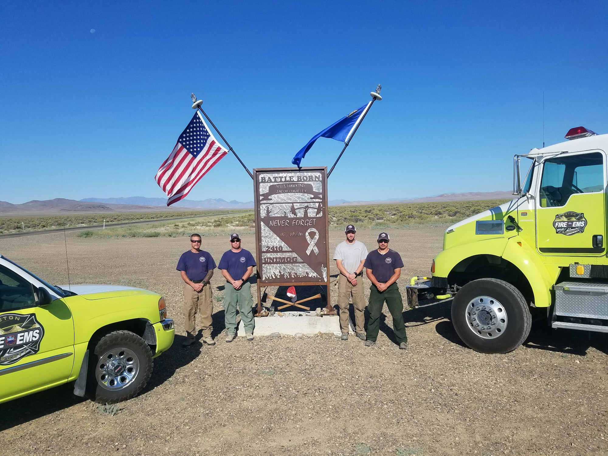 Four firefighters by a memorial, vehicles, US & state flags, desert landscape.