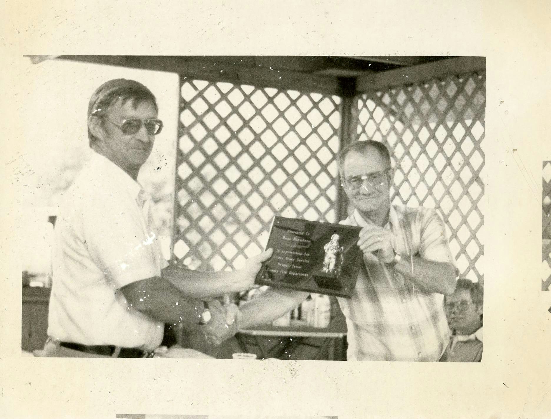 Two men shaking hands, one receiving a plaque, in a room with a lattice backdrop. Vintage photo aesthetic.