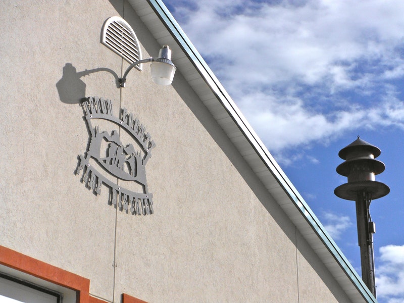 Wall-mounted outdoor light casting a shadow of a bell and text next to a siren on a building under a blue sky.