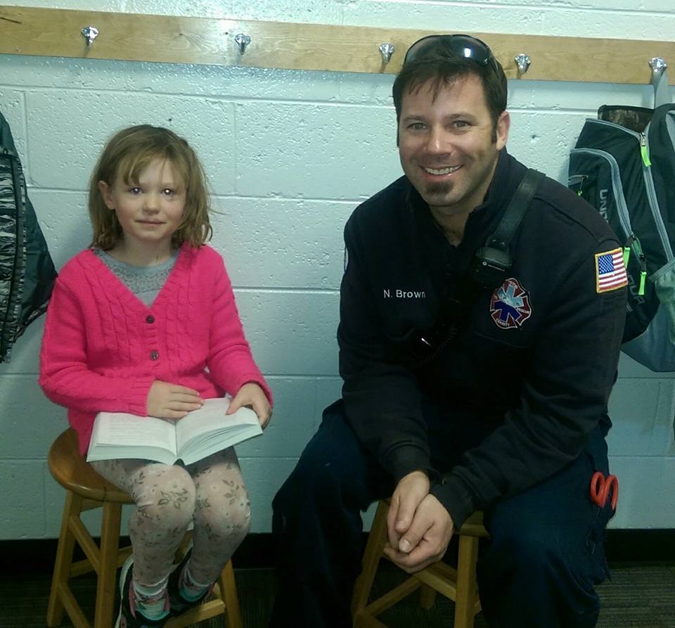 A young girl reading a book next to a smiling man in a paramedic uniform, both sitting on stools.
