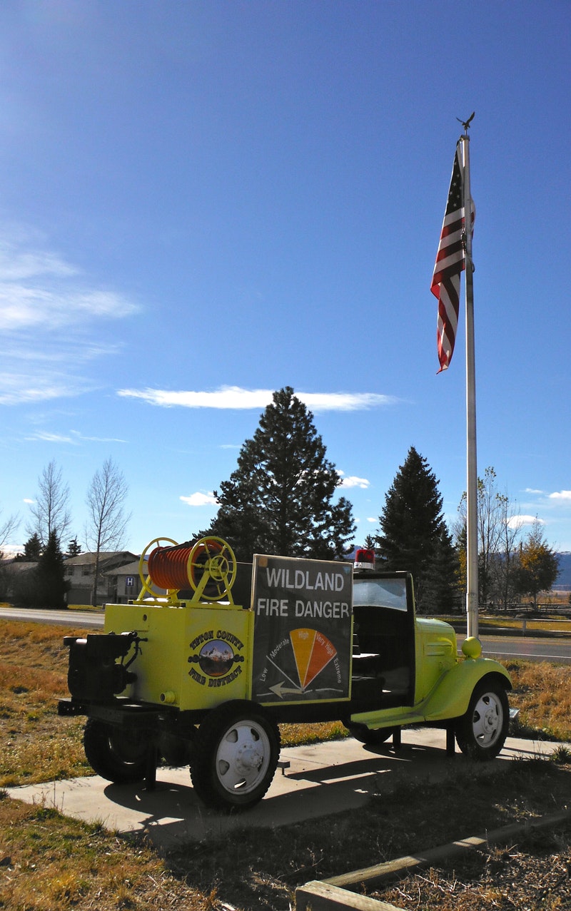 A vintage yellow fire danger sign vehicle and an American flag on a clear day.
