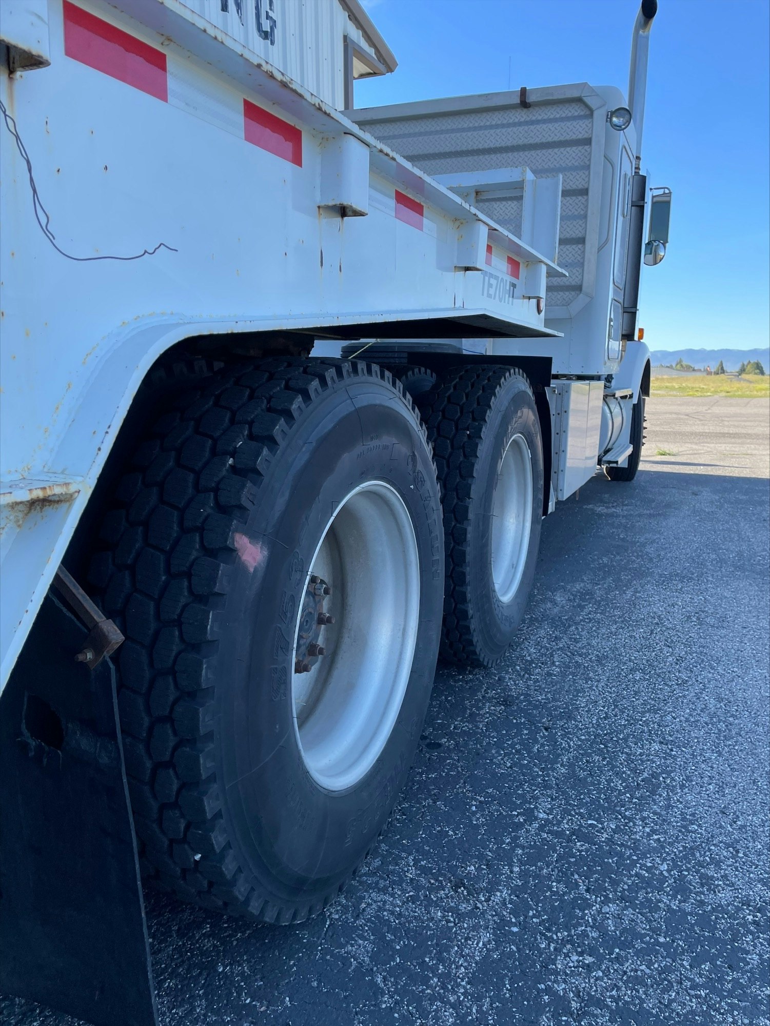 White semi-truck with large tires, part of trailer visible, clear blue sky.
