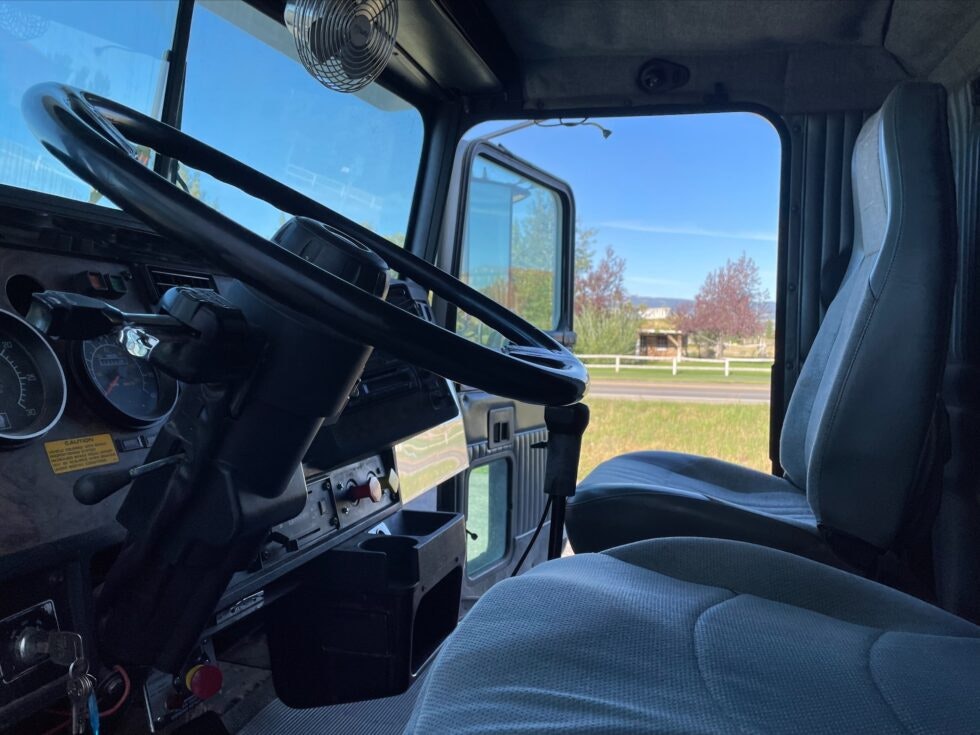 Interior of a truck cabin with steering wheel, dashboard, and seats, parked by a roadside.