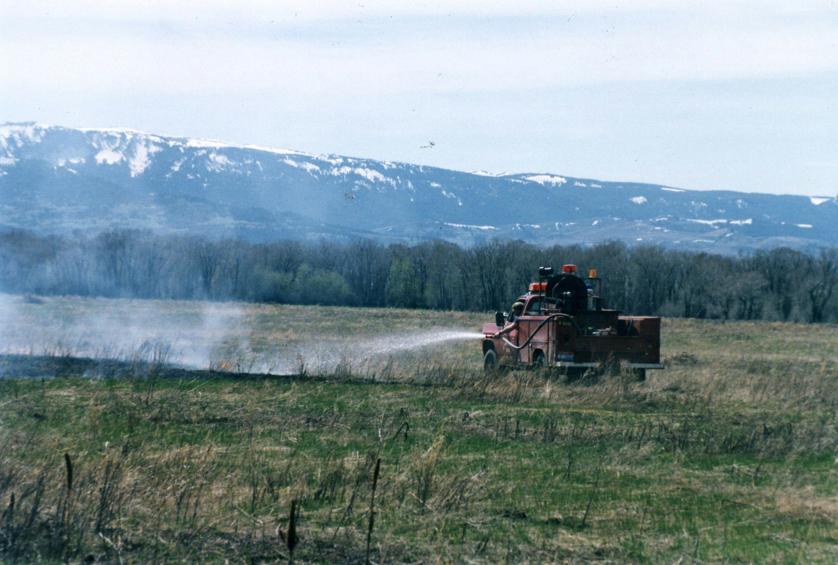 A fire truck spraying water near a small fire, in a field with a mountainous backdrop.