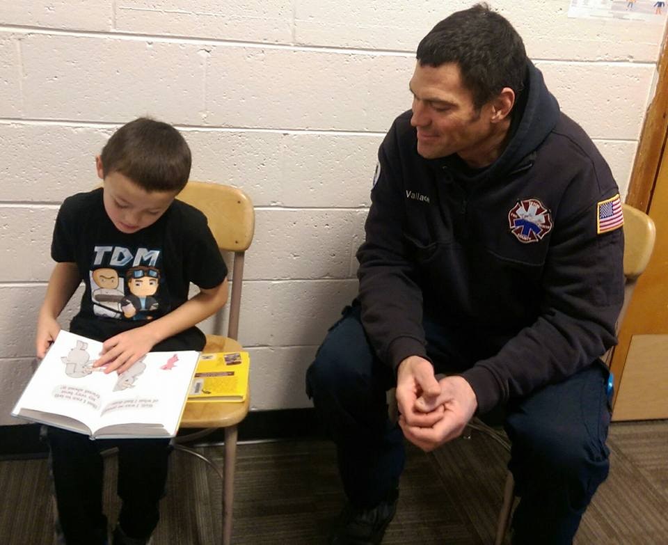 A child is reading a book sitting next to a firefighter.