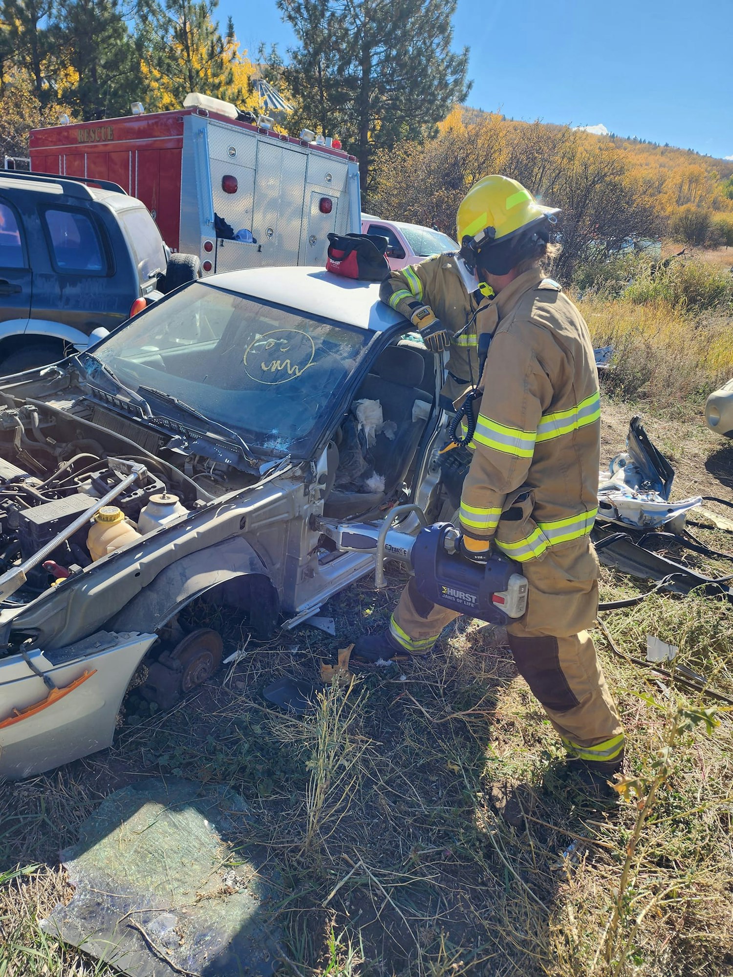 A firefighter stands by a severely damaged car with rescue equipment, in daylight with foliage in the background.