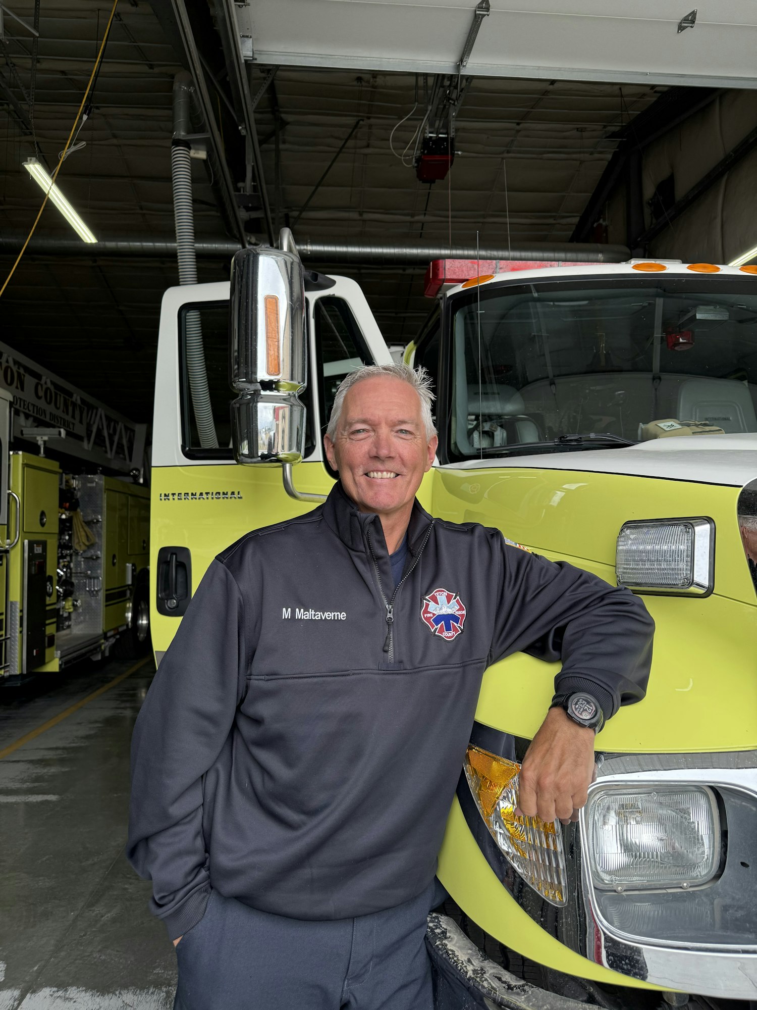 A person in a uniform stands smiling in a garage, leaning on a yellow fire truck.